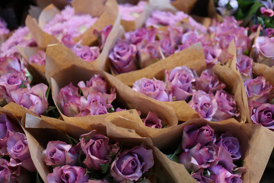 A close-up view of numerous bouquets of fresh, light purple roses nestled in brown craft wrapping paper, arranged in a floral display. The roses feature velvety petals with subtle color variations fro